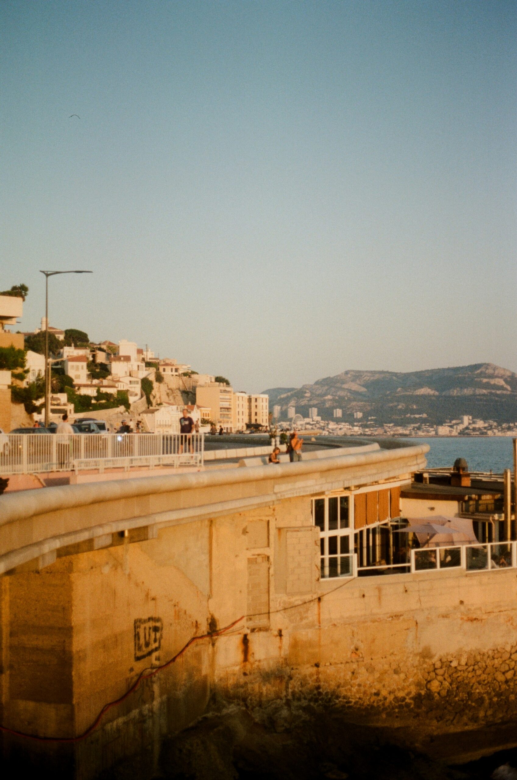 A coastal promenade above a rocky shoreline at golden hour, with travelers walking along the water