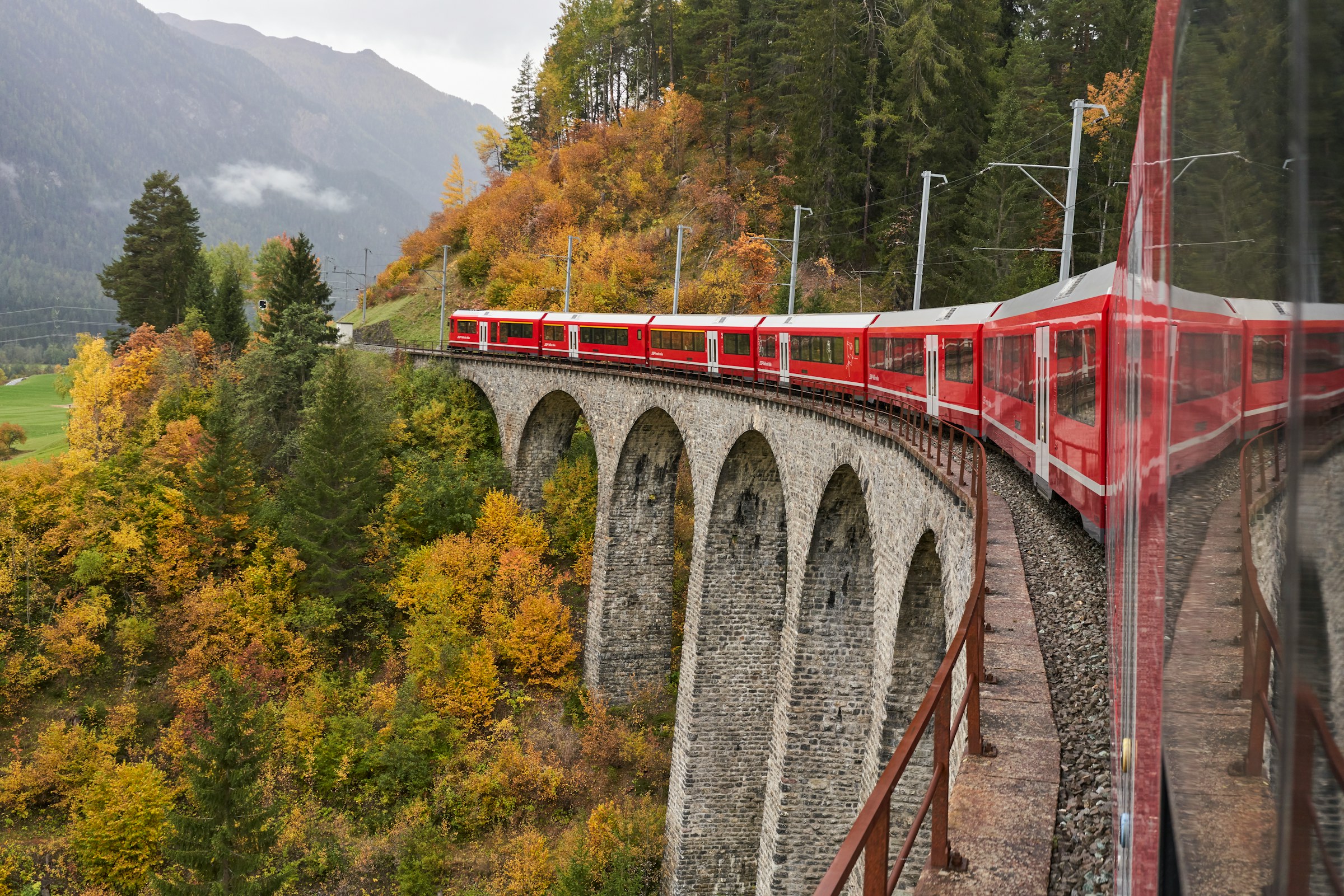 A red mountain train crossing a curved stone viaduct through autumn forest in the Swiss Alps