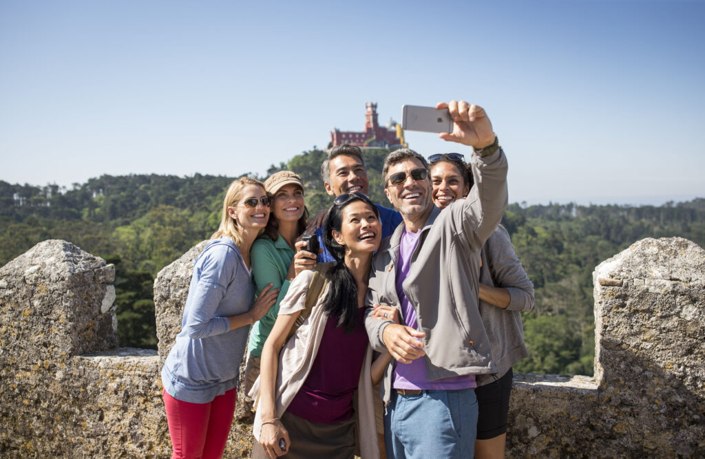 Smiling Globus travelers take a group selfie on castle walls in Sintra, Portugal, with Pena Palace on the hilltop behind them